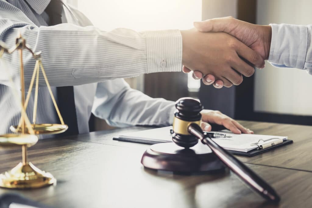 A close-up of two people shaking hands over a wooden desk. A gavel and a set of brass scales of justice are visible on the desk, along with a clipboard with papers. Soft natural light comes from the background.