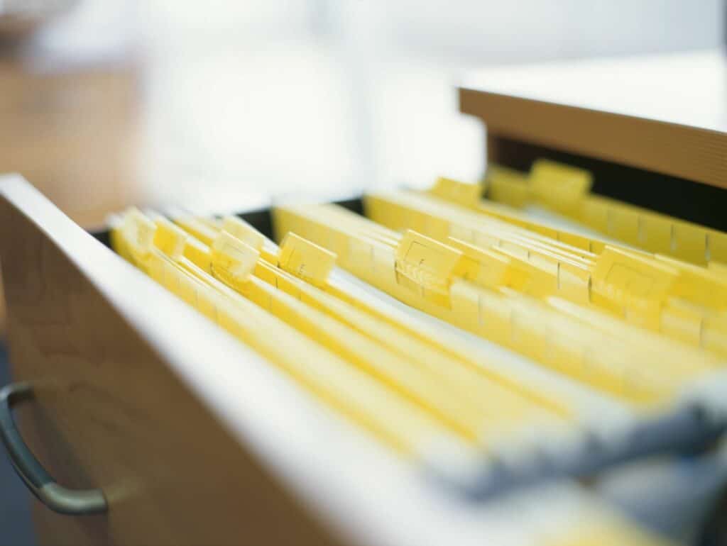 An open drawer filled with hanging file folders, featuring yellow folders neatly organized with labeled tabs. The background is softly blurred, suggesting an office environment with natural light.