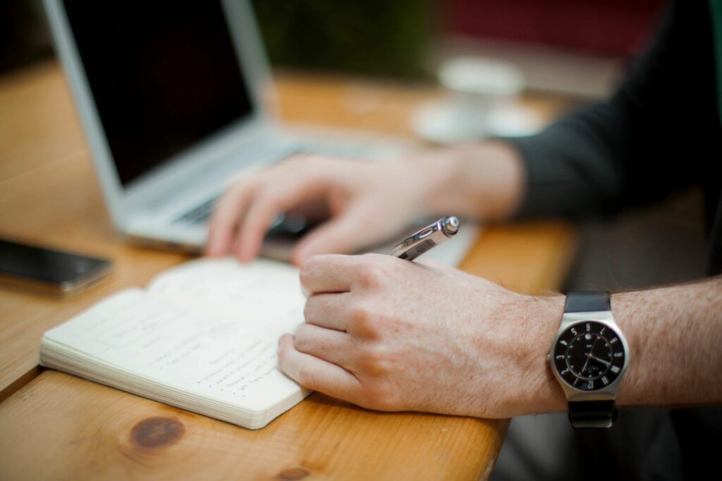 A close-up view of a person's hand holding a pen, writing in a notebook on a wooden table. In the background, there is a laptop and a cup of coffee, indicating a workspace setting.