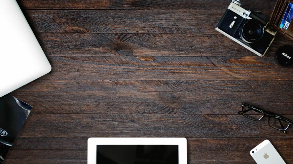 A flat lay view of a wooden desk with a closed laptop, an iPad, a pair of glasses, a vintage camera, a smartphone, and a black notebook aligned around the edges. The surface features dark wooden texture, enhancing the workspace aesthetic.