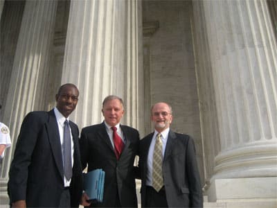 Three men pose together in front of large classical columns of a building. The man on the left wears a dark suit, the central man is in a dark suit with a red tie, and the man on the right dons a light suit with a patterned tie.