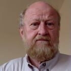 A man with a gray beard and bald head wearing a light-colored button-up shirt is facing the camera. He is positioned against a neutral-colored background.