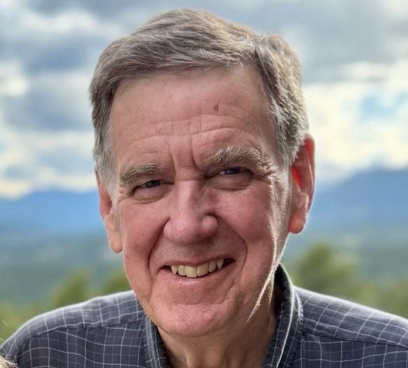 A smiling older man in a dark checkered shirt, shown in a close-up portrait with mountains and cloudy sky in the background.