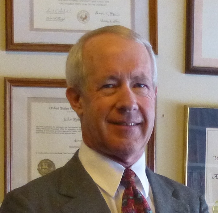 A portrait of an older man in a suit and patterned tie standing in front of framed certificates on a wall.